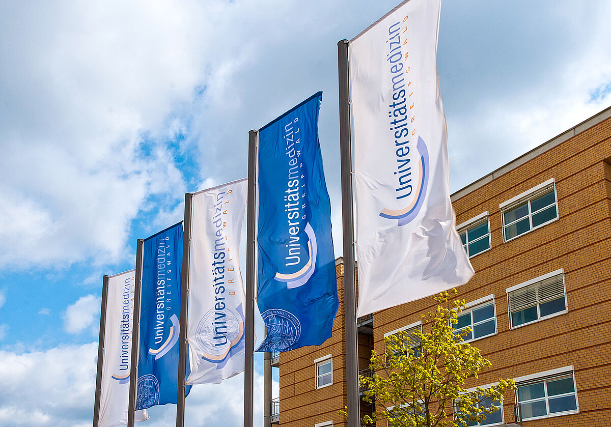 Symbolbild: Drei Flaggen mit dem Logo der Universitätsmedizin Greifswald vor eine Häuserfront.