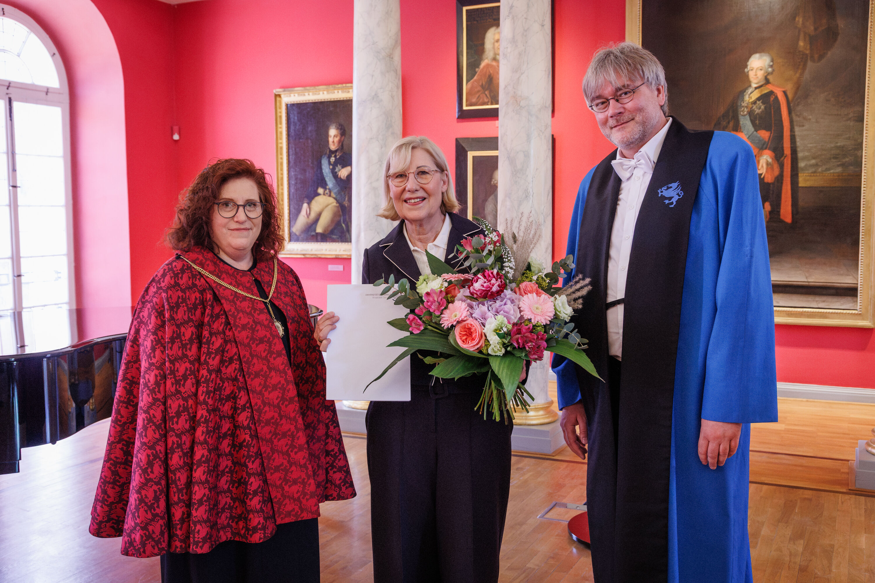 Ehrenpromotion Ursula Gather Drei Menschen stehen in einem eleganten Raum, eine Frau wurde geehrt und hält einen Blumenstrauß in der Hand.