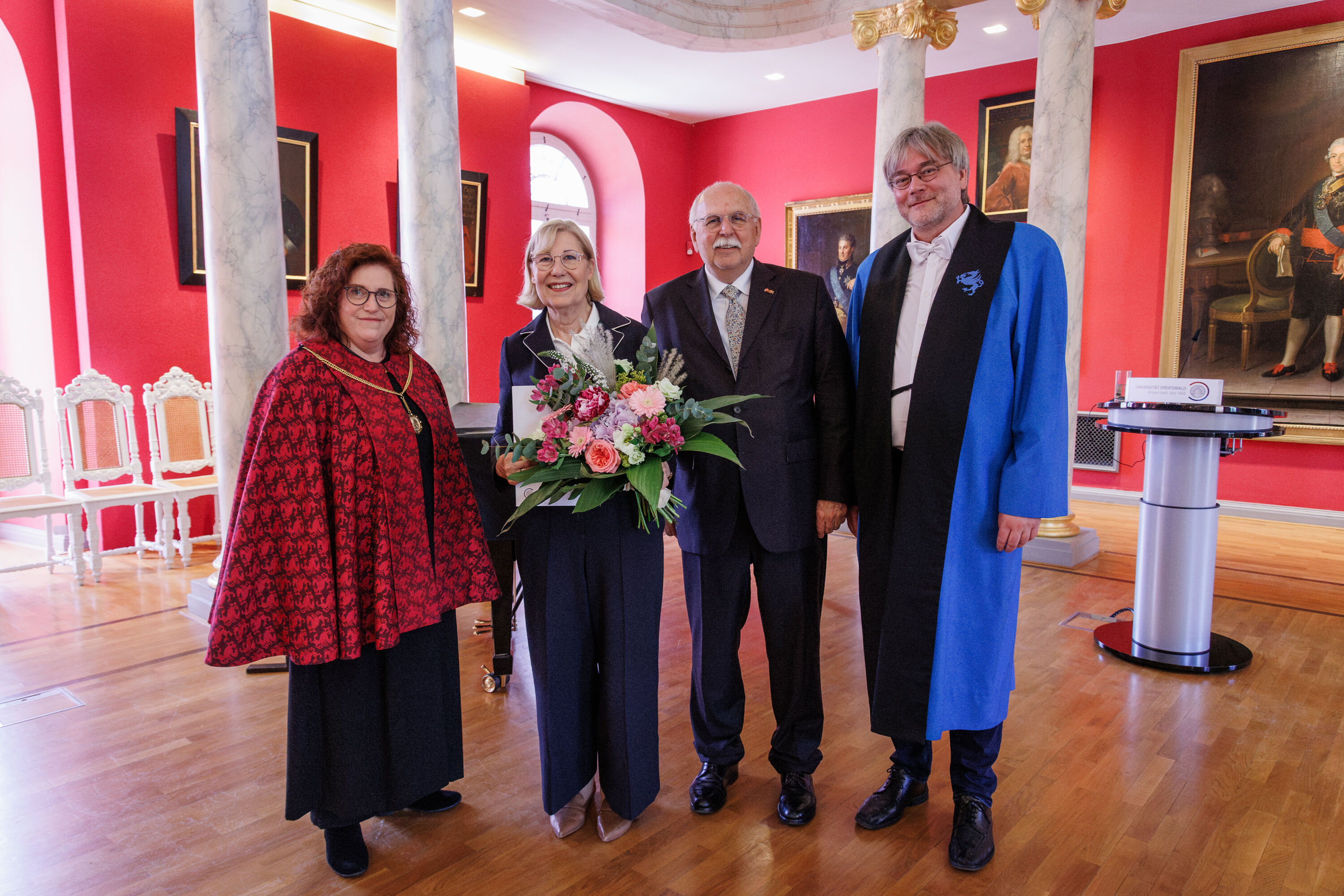 Ehrenpromotion Ursula Gather Vier Menschen stehen in einem eleganten Raum, eine Frau wurde geehrt und hält einen Blumenstrauß in der Hand.
