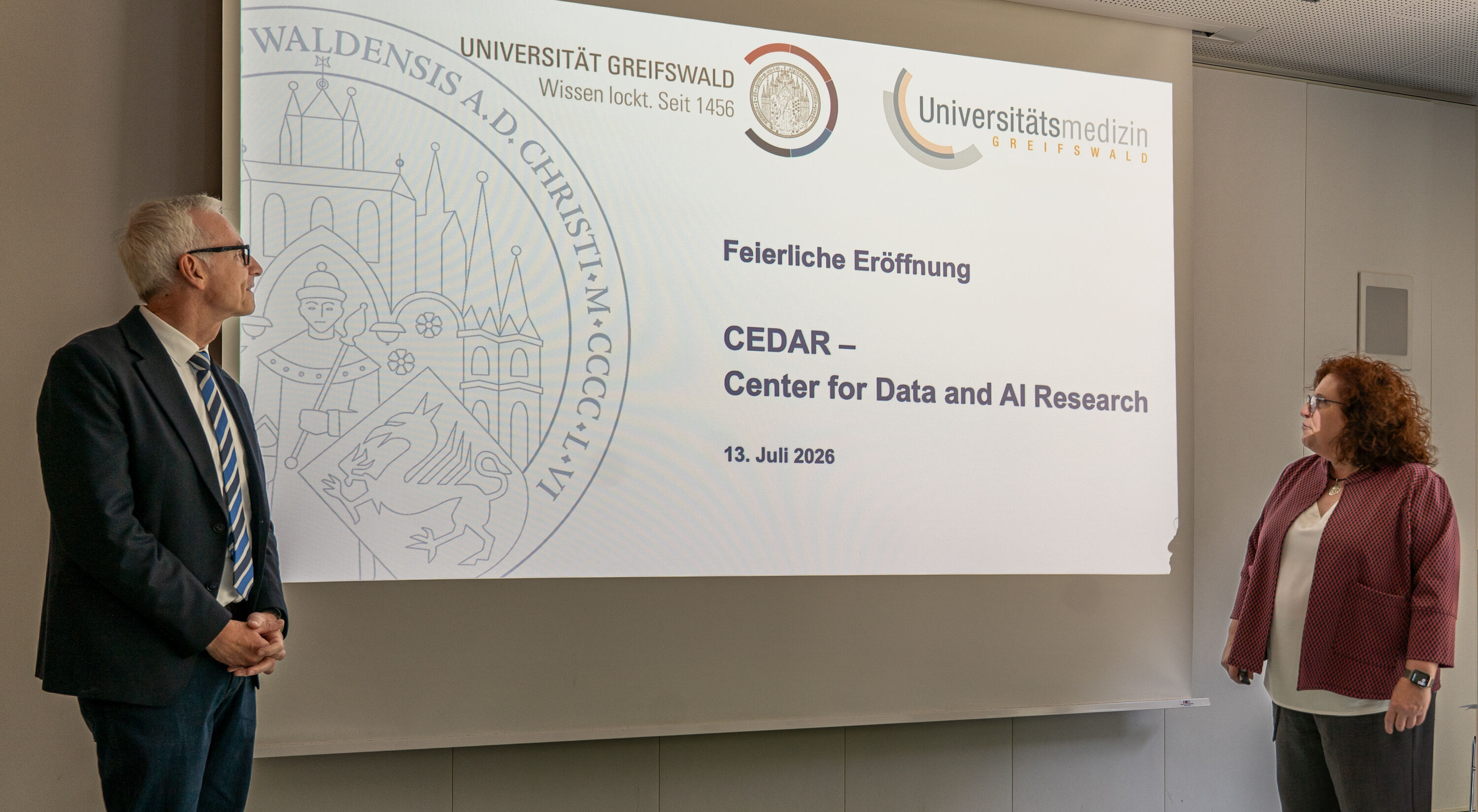 Prof. Dr. Karlhans Endlich und Prof. Dr. Katharina Riedel eröffnen das CEDAR Zwei Personen, ein mann und eine Frau, stehen vor einer Präsenation und halten eine Ansprache.
