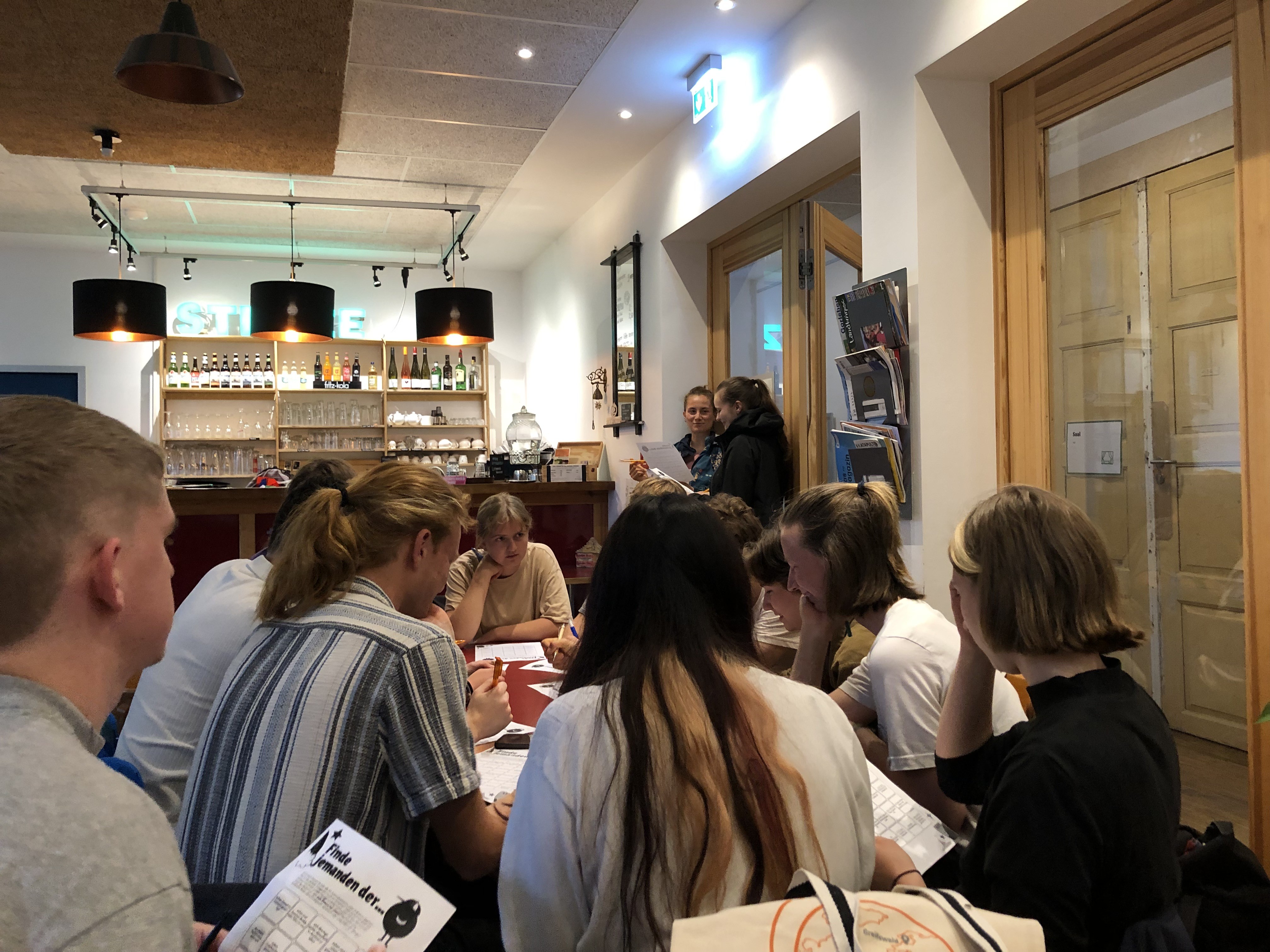 Mehrere Personen sitzen in einem Restaurant rund um einen Tisch und unterhalten sich. Im Hintergrund ist die Bar zu sehen.