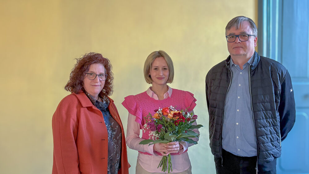 Drei Personen stehen in einer Reihe, die in der Mitte hält einen Blumenstrauß