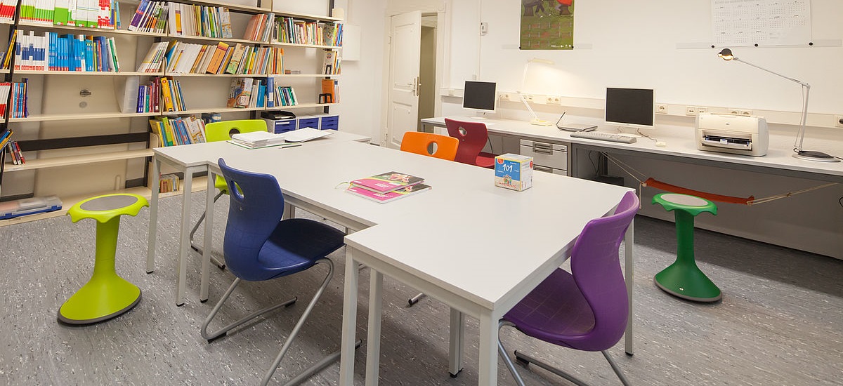Blick in einen Seminarraum: Ein Bücherregal steht links an der Wand. Im Hintergrund rechts stehen Tische mit zwei Monitoren sowie einem Drucker. Im Vordergrund steht ein Tisch, um den Hocker und Stühle gestellt wurden.