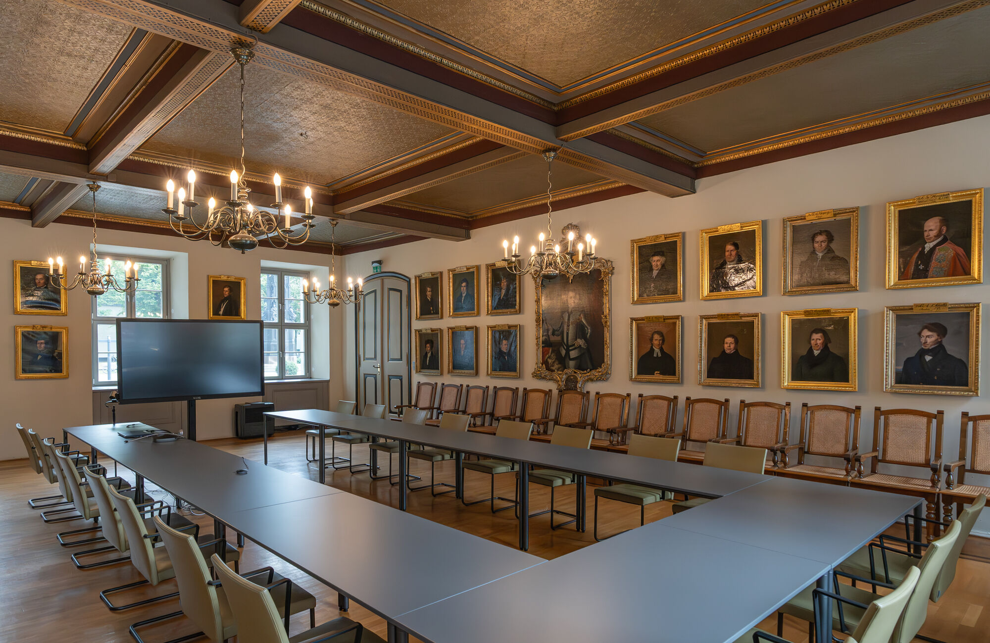 Blick in den Konzilsaal In der Mitte stehen in U-Form arrangierte Tische. Im Hintergrund hängen die golden gerahmten Portraits ehemaliger Rektor*innen. Die Decke ist mit einer schönen Holzfassade geschmückt. Es sieht festlich aus.