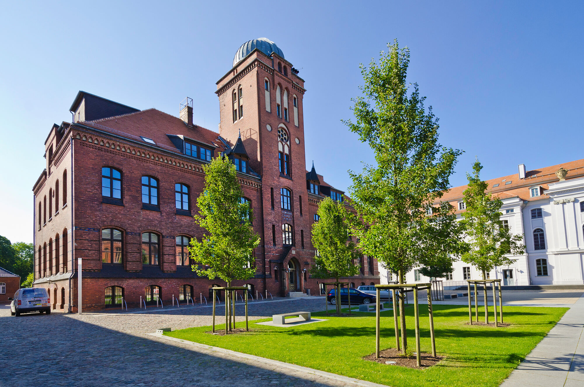 Backsteinernes Gebäude der Alten Physik Greifswald. Im Vordergrund befinden sich sonnenbeschienene junge Bäume. Die Sternwarte mit ihrer charakteristischen Kuppel auf dem Dach des Gebäudes stellt sich vor blauem Himmel dar. 