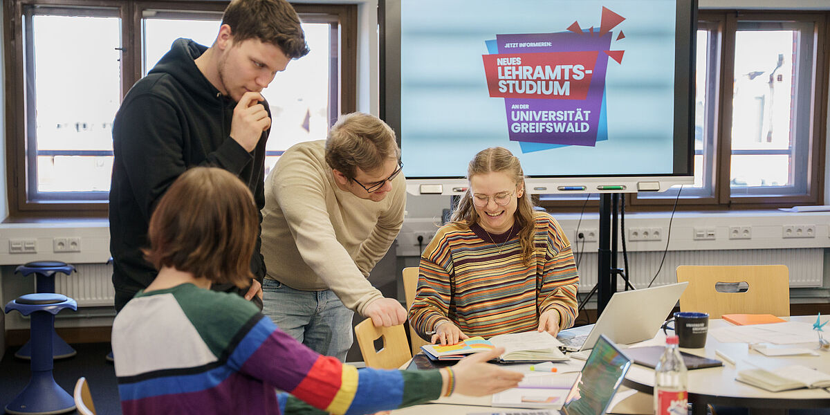 Es befinden sich vier Studierende in einem Raum an der Uni, welche gemeinsam an einem großen Tisch studieren und in ihre Bücher gucken. Im Hintergrund ist ein großer Bildschirm mit der Aufschrift "Neues Lehramtsstudium".