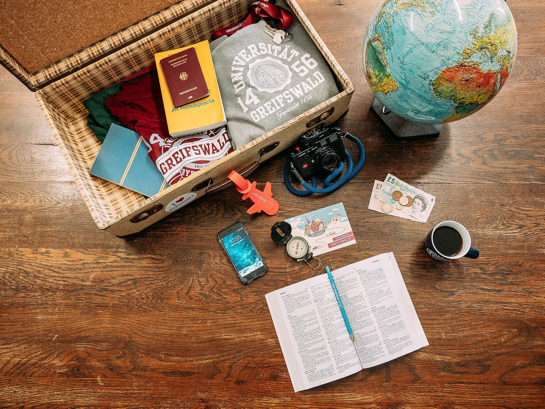 Koffer / Stillleben / International Office Auf einem Tisch liegt ein geöffneter Koffer, in ihm liegen ein grauer Hoodie, ein rotes T-Shirt, ein gelbes Wörterbuch, ein Schlüssel mit rotem Schlüsselband sowie ein Pass. Neben dem Koffer steht ein Globus. Vor dem Koffer liegt eine Kamera, ein Smartphone, ein Kompass sowie Geld und ein aufgeschlagenes Wörterbuch. Daneben steht eine Tasse mit Kaffee.