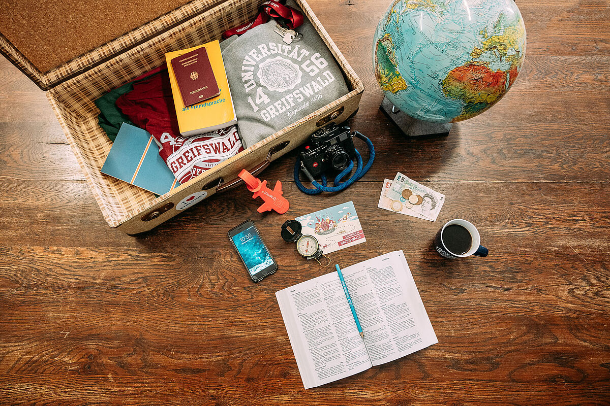 Auf einem Tisch liegt ein geöffneter Koffer, in ihm liegen ein grauer Hoodie, ein rotes T-Shirt, ein gelbes Wörterbuch, ein Schlüssel mit rotem Schlüsselband sowie ein Pass. Neben dem Koffer steht ein Globus. Vor dem Koffer liegt eine Kamera, ein Smartphone, ein Kompass sowie Geld und ein aufgeschlagenes Wörterbuch. Daneben steht eine Tasse mit Kaffee. 