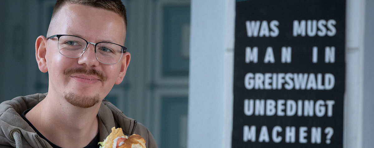 Ein Campusspezialist hält ein Fischbrötchen in der Hand und sieht dabei sehr glücklich aus. Neben ihm ist eine Tafel zu sehen mit der Aufschrift "Was muss man in Greifswald unbedingt machen?"