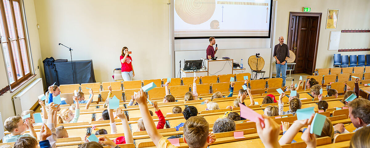 Symboldbild Kinder- und JugendUNI 2025 Viele Kinder sitzen in einem Hörsaal der Universität Greifswald und heben ihre Finger, um Fragen zu beantworten, die die Dozierenden stellen, die vorne stehen und einen Vortrag halten.