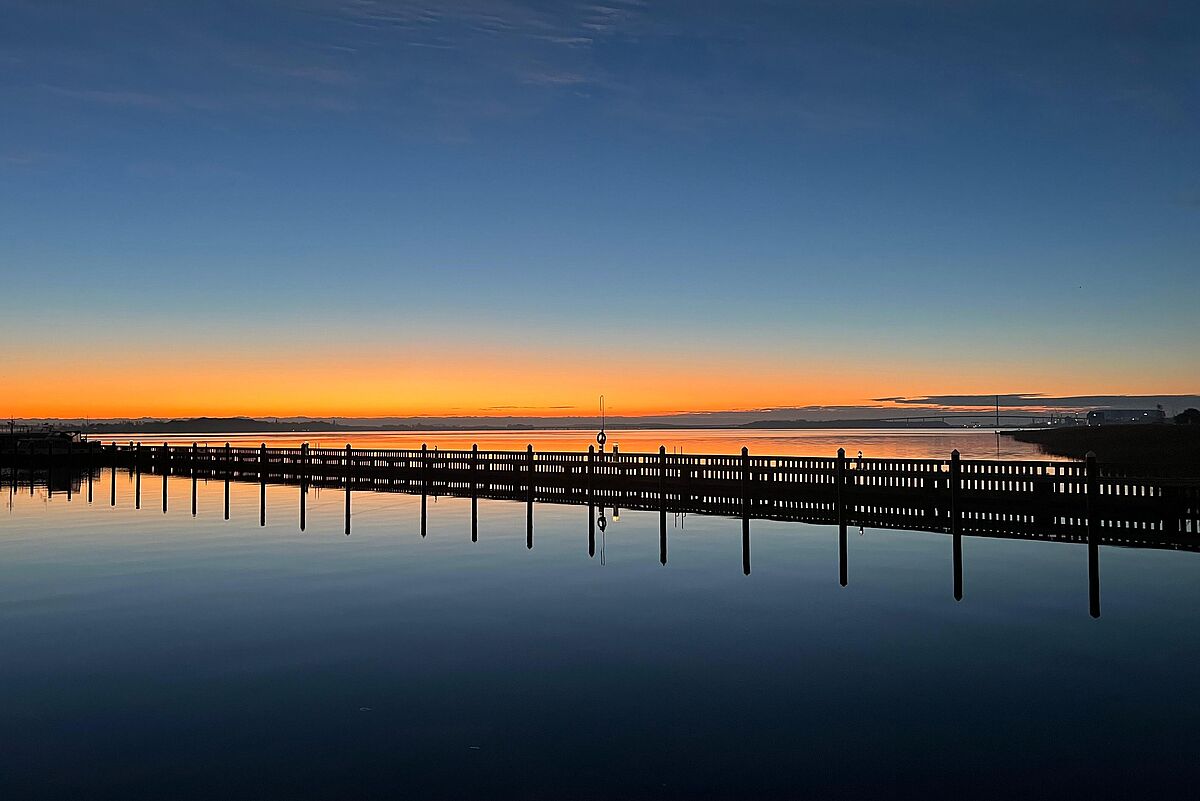 Stralsund Sonnenaufgang Eine Brücke über windstillem Wasser, in dem sich das orange und dunkelblaue Licht des Sonnenaufgangs spiegelt.
