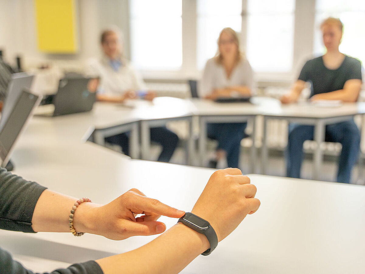 Im Vordergrund wird eine Smartwatch an einem Handgelenk bedient, während im Hintergrund unscharf vier Personen an Tischen in einem hellen Raum sitzen.