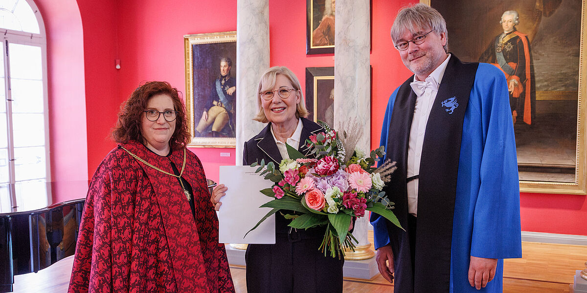Ehrenpromotion Ursula Gather Drei Menschen stehen in einem eleganten Raum, eine Frau wurde geehrt und hält einen Blumenstrauß in der Hand.