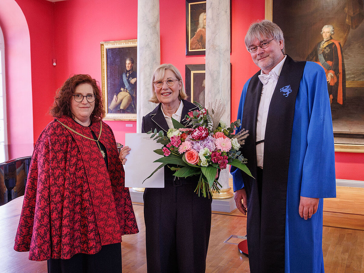 Drei Menschen stehen in einem eleganten Raum, eine Frau wurde geehrt und hält einen Blumenstrauß in der Hand.
