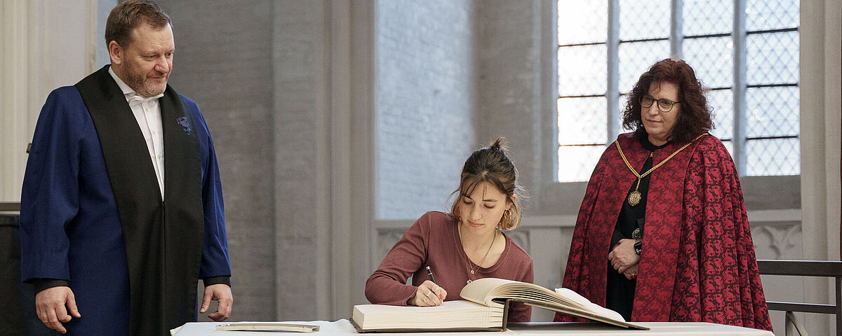 Eine Person sitzt an einem Tisch und  schreibt in ein großes Buch. Links und rechts neben ihr stehen der Dekan und die Rektorin der Universität.