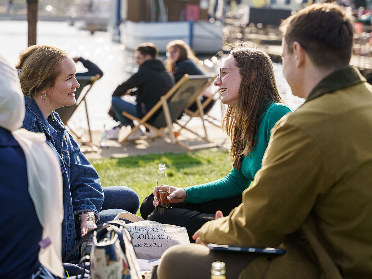 Eine Gruppe studierener sitzt ausgelassen bei Sonnenschein am Hafen und unterhält sich. Im Hintergrund schimmert das Wasser.