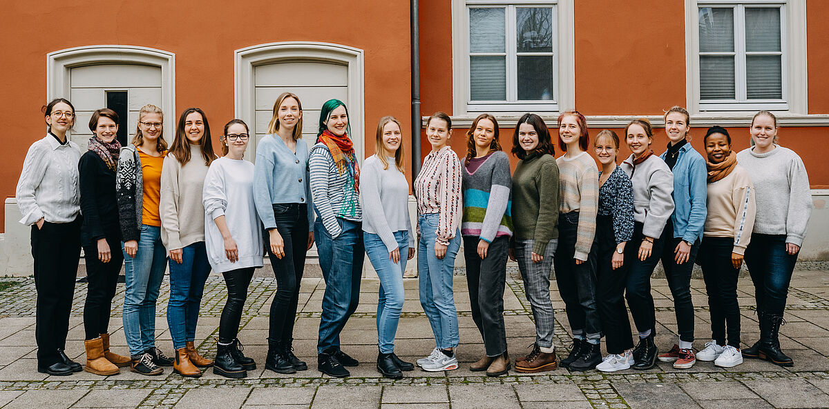 17 Frauen stehen in einer Reihe nebeneinander, alle lächeln in die Kamera.