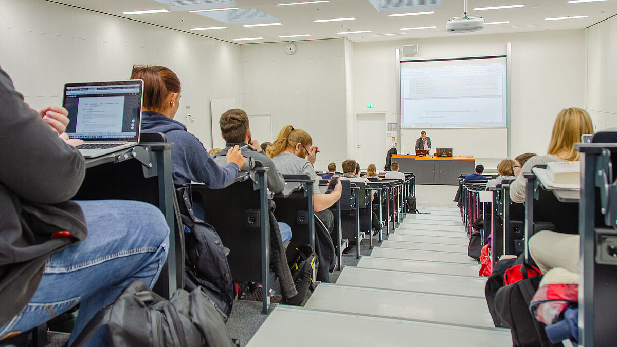 Vorlesung im Hörsaal; Studierende machen sich Notizen während einer Vorlesung. Das Foto ist in einem Gang zwischen den Sitzreihen aufgenommen und zeigt an dessen Ende den Dozierenden.