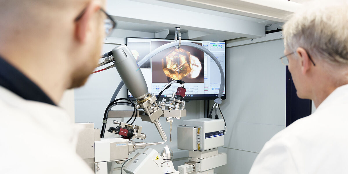 Forschungsbilder Biochemie Von hinten sieht man unscharf zwei Männer in weßen Kitteln, die auf einen Bildschirm schauen, vor dem sich eine technische Gerätschaft zum Vergrößern von Materialien befindet.