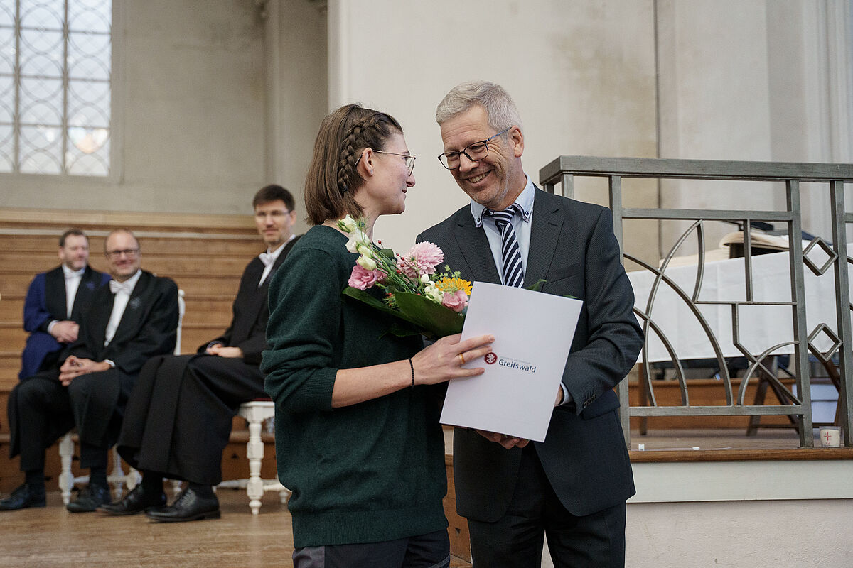 The winner of the Mayor's Prize and the Mayor himself stand in Greifswald Cathedral and shake hands, while the Mayor presents the young woman with flowers and a certificate. Members of the rectorate sit at the back of the church and watch.
