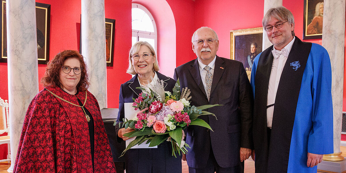 Ehrenpromotion Ursula Gather Vier Menschen stehen in einem eleganten Raum, eine Frau wurde geehrt und hält einen Blumenstrauß in der Hand.