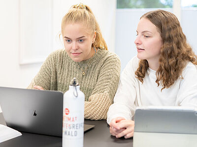 Zwei Studentinnen sitzen nebeneinander an ihren Notebooks und tauschen sich aus.