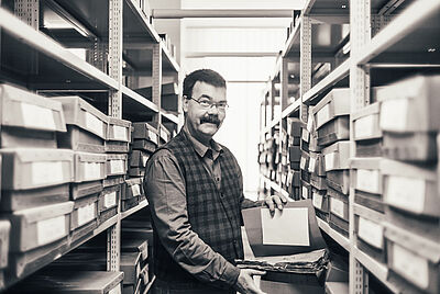 Auf dem Foto ist der Verstrobene bei der Arbeit im Archiv zu sehen. Er steht zwischen den Regalen und blättert in einer Akte und lächelt.