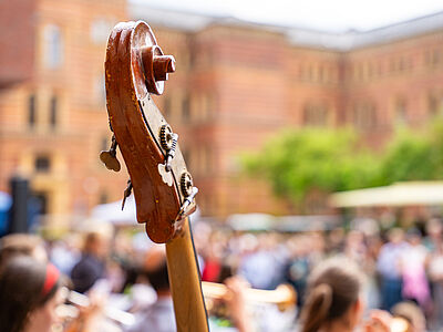 Der Hals einer Violine ist im Vodergrund zu sehen. Im Hintergrund sitzen schemenhaft Menschen, die dem sommerlichen Konzert beiwohnen.