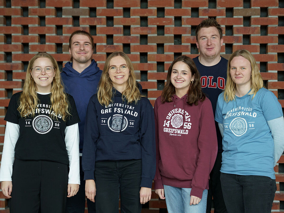 Guppenbild Campusspezialist*innen im Foyer der Uni vor einer steinernden Wand. Zu sehen sind sechs Personen, vorne stehen vier Campusspezialistinnen, sie tragen Uni Greifswald T-Shirts und dahinter stehen zwei Campusspezialisten. Alle lächeln freundlich in die Kamera.