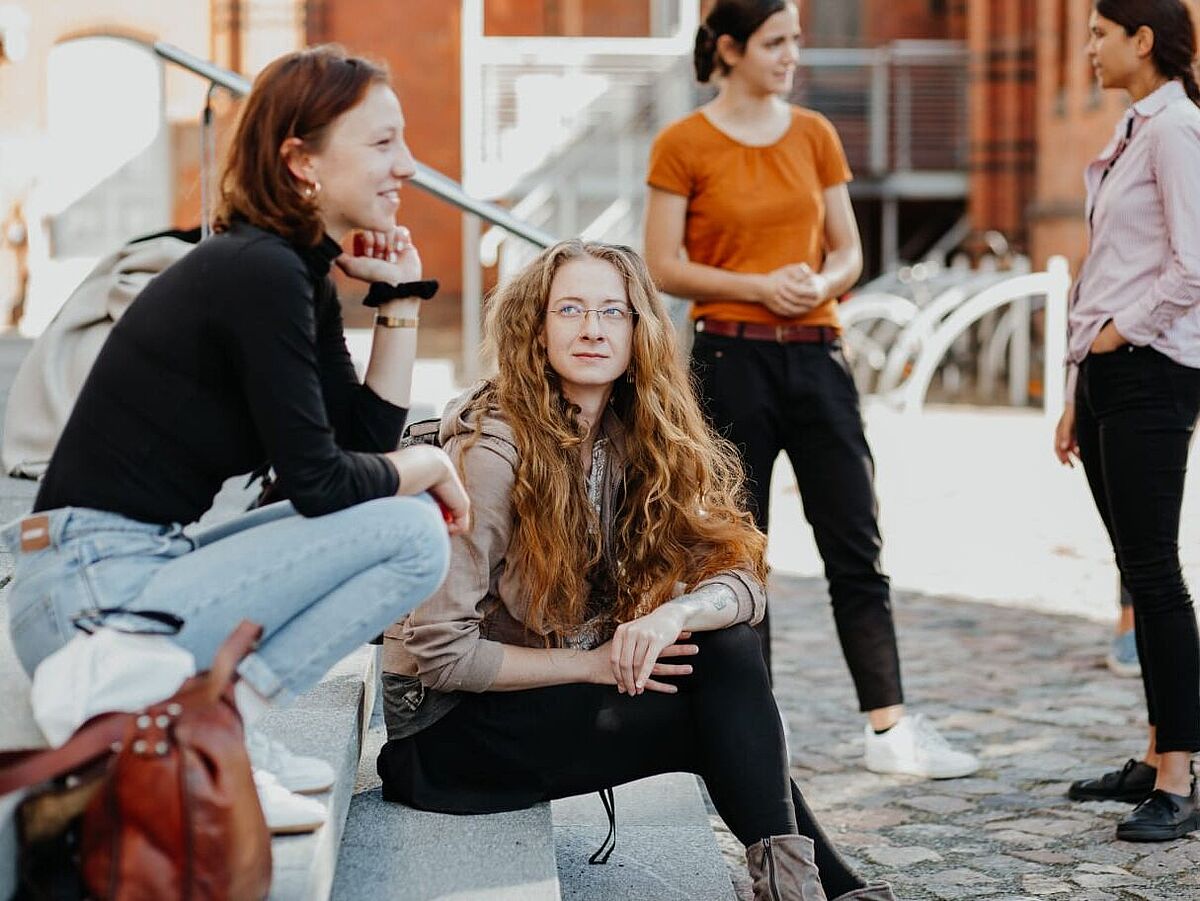 Junge Frauen sitzen auf der Treppe vor dem Unihauptgebäude und sprechen miteinander..