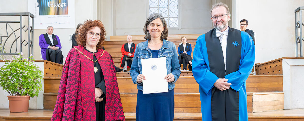 Bild von Franziska Tanneberger im festlich geschmückten Dom vor der Holztreppe. Sie trägt ein dunkelblaues Kleid und eine hellblaue Jeansjacke und hält ihr Zertifikat über ihre Habilitation in der Hand. Zu ihrer rechten Seite steht die Rektorin und zu ihrer linken Seite der Dekan der Mathematisch-Naturwissenschaftlichen Faktultät. 