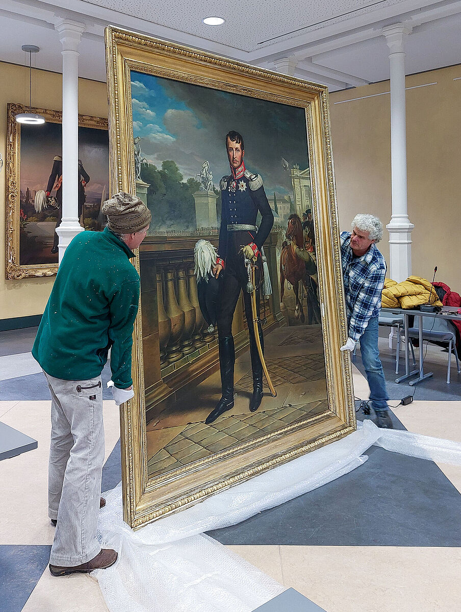 Das Gemälde kehrt zurück in den Konferenzsaal der Universität. Es ist zu sehen, wie zwei Mitarbeitende der Resaturierungswekrstatt das Gemälde auspacken.