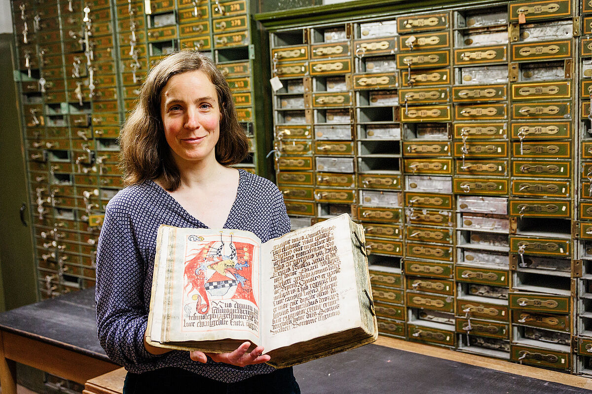 Archivleiterin Dr. Elisabeth Heigl Eine Frau lächelt in die Kamera während sie in einem Archiv steht und ein altes Buch in die Kamera zeigt.