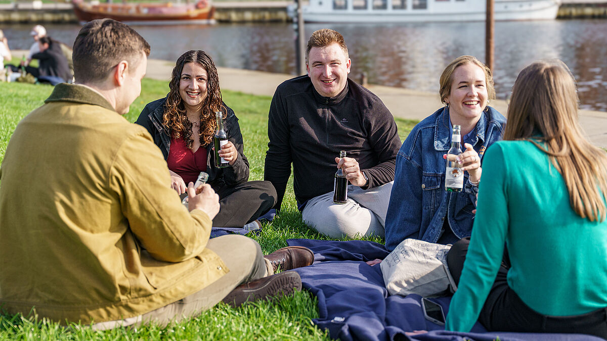 Fünf Studierende sitzen auf dem Rasen am Ryck, unterhalten sich und halten Erfrischungsgetränke in der Hand.