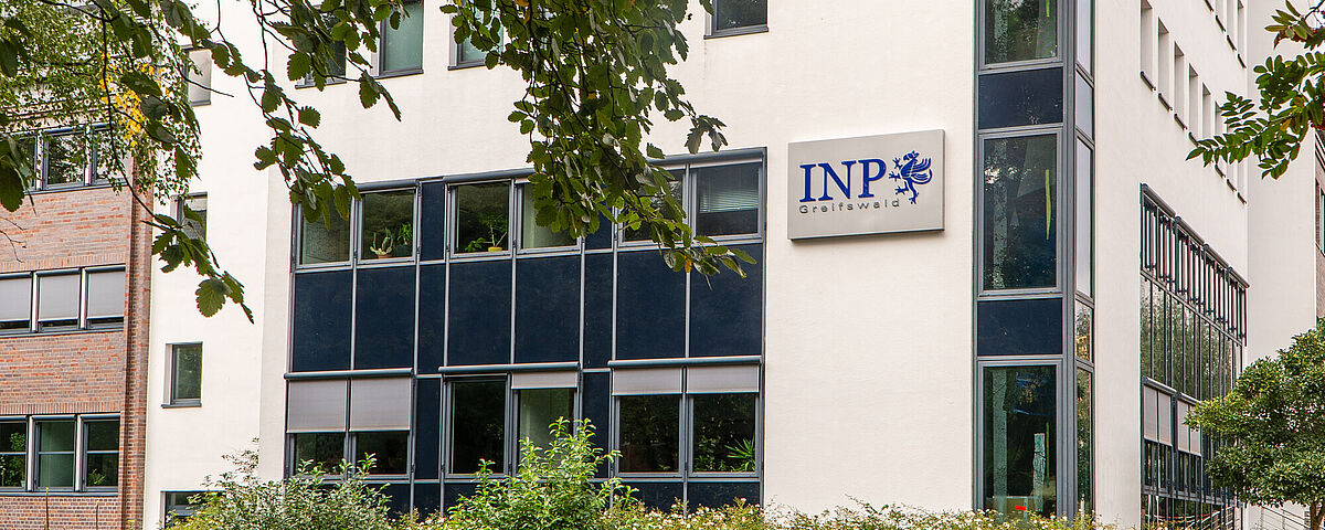 Ein weißes Gebäude mit einem Schild, auf dem INP - Leibniz-Institut für Plasmaforschung steht und von grünen Bäumen gerahmt ist.