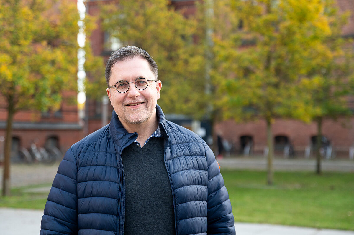 Prof. Dr. Kilian Heck vor Bäumen und Gebäude, ein heer in blauer Jacke und Brille