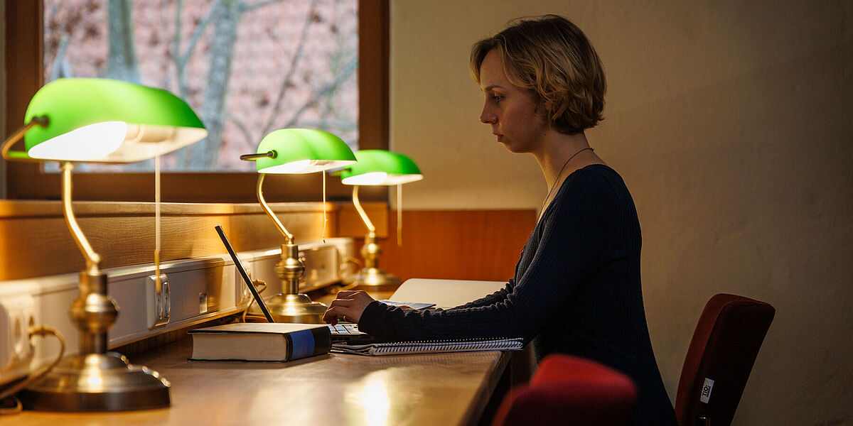 Im Lesesaal einer Bibliothek liest eine Studentin in einem Buch. Auf den Tischen leuchten grüne Tischlampen.