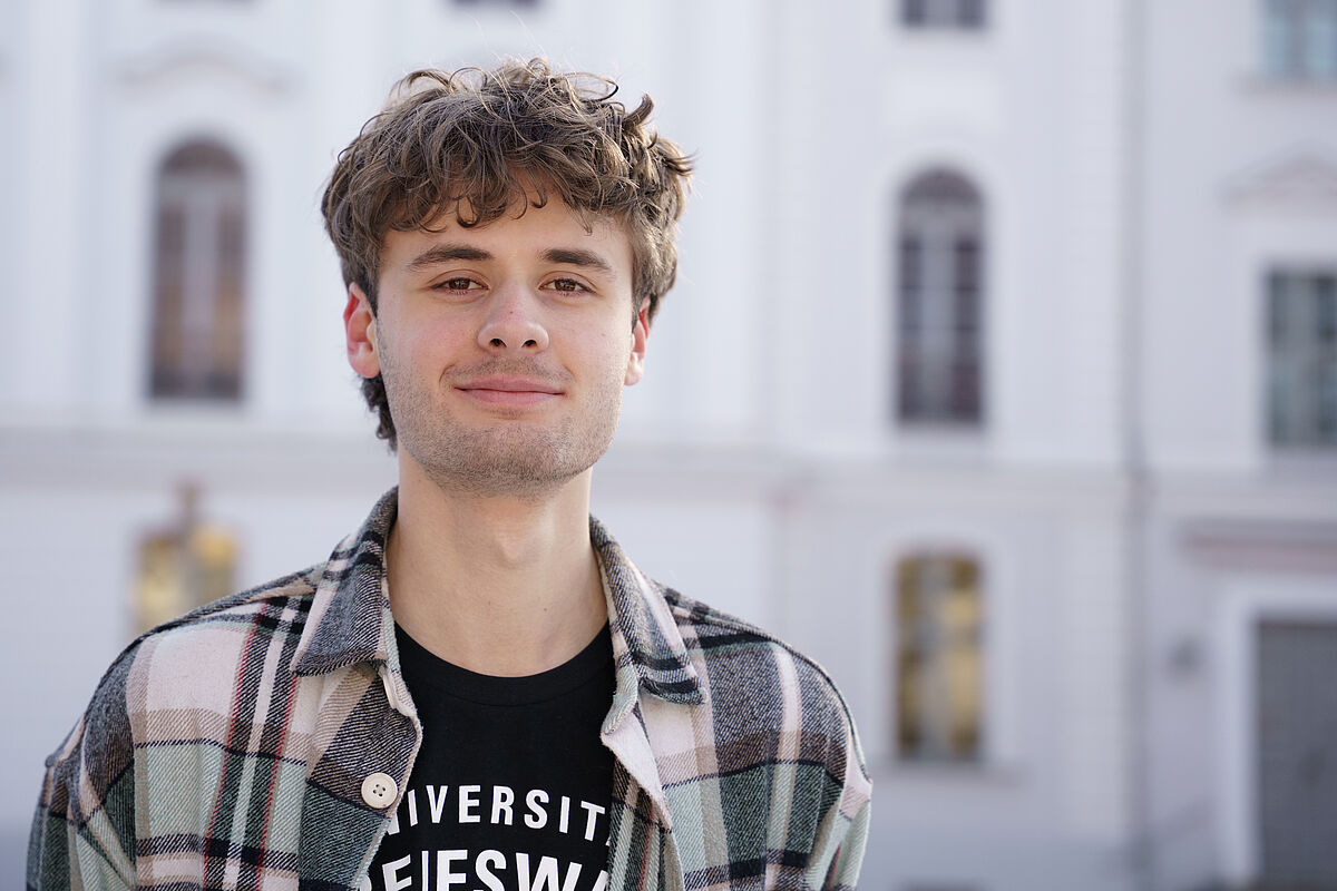 Ein Porträt von Eric Recknagel. Er lächelt in die Kamera und steht vor dem Universitätshauptgebäude.