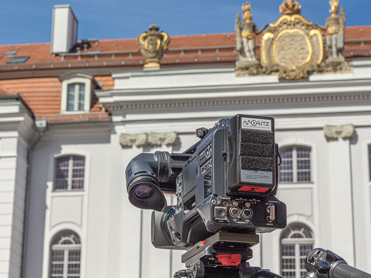 Stock Image Science Communication In the foreground is a professional film camera mounted on a tripod; in the background is the university's main building with its white Baroque facade featuring ornate elements and red roof tiles.