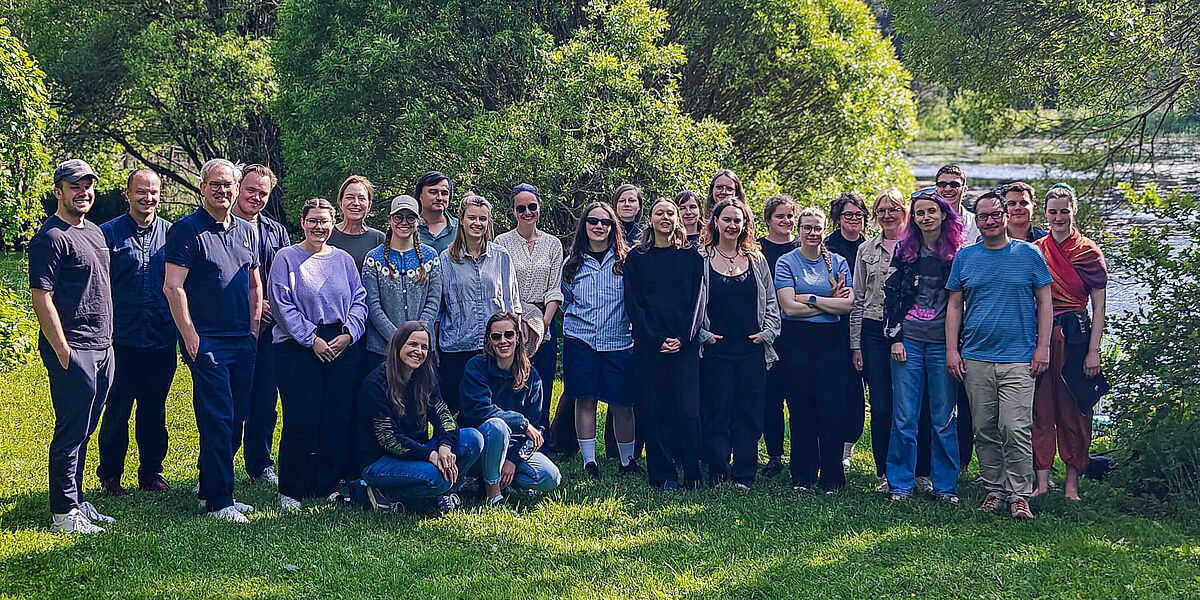 Eine große Gruppe nett lächelnder Menschen steht auf einer grünen Wiese vor grünen Bäumen.