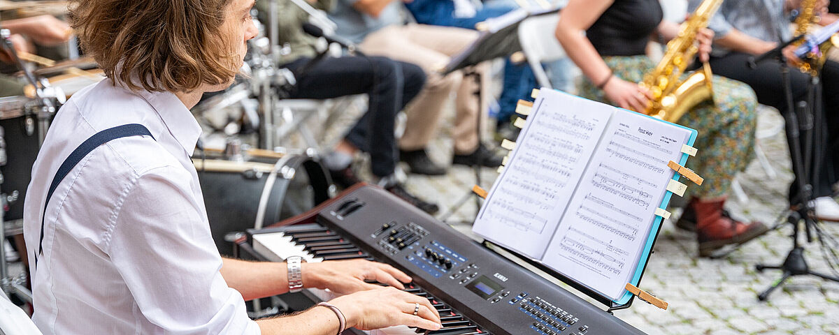 Ein Mann sitzt spielend am E-Piano. Im Hintergrund sitzen Menschen mit Blasinstrumenten.