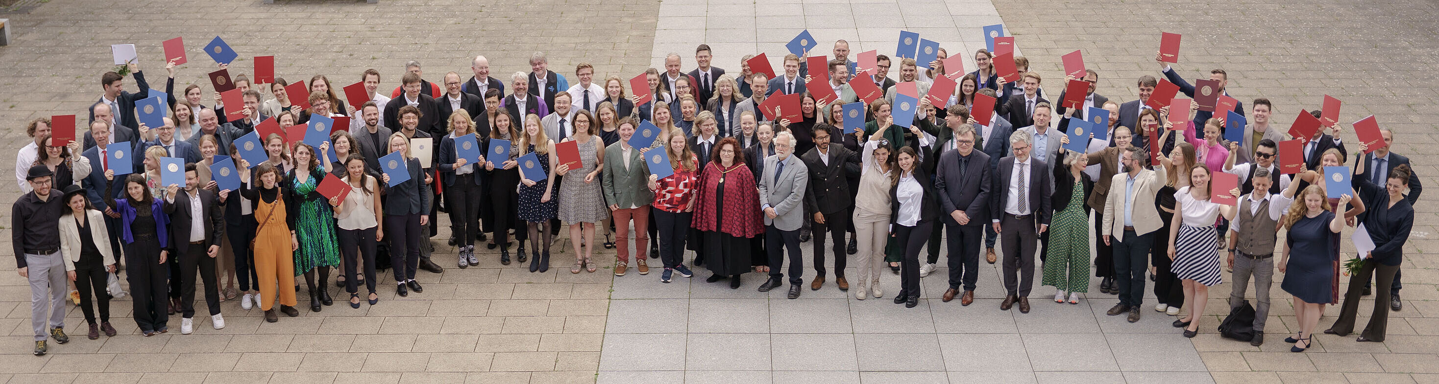 Viele Menschen stehen für ein Gruppenbild im Innenhof einer alten Universität und halten Abschlussurkunden in die Luft.