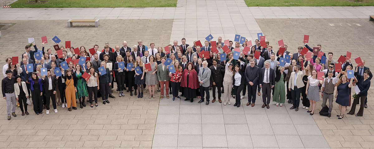 Verleihung akademishcer Grade Viele Menschen stehen für ein Gruppenbild im Innenhof einer alten Universität und halten Abschlussurkunden in die Luft.