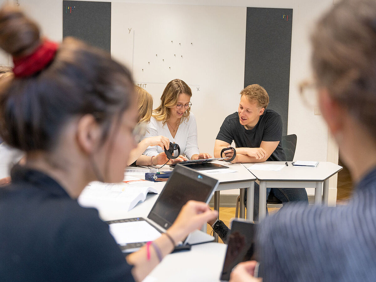 Im Meeting: Eine Gruppe von Menschen sitzt an weißen Tischen in einem Raum und arbeitet gemeinsam mit Tablets und VR-Controllern. Der Fokus liegt auf zwei Personen in der Mitte, die lächeln und ein Gerät bedienen.