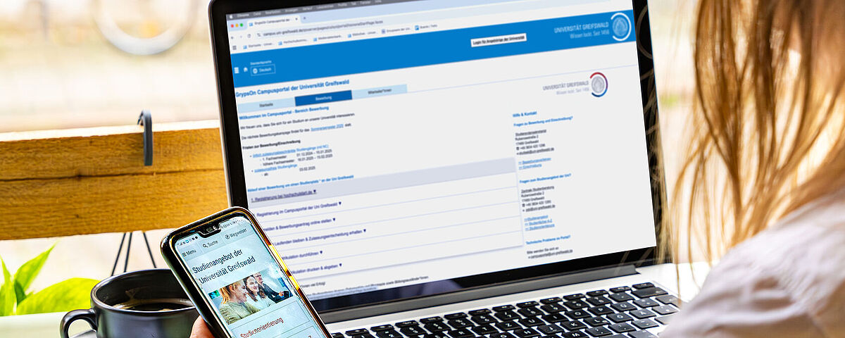 Eine Person sitzt vor ihrem geöffneten Laptop und schaut zusätzlich in ihr Smartphone, wo die Seite zur Studienorientierung geöffnet ist.
