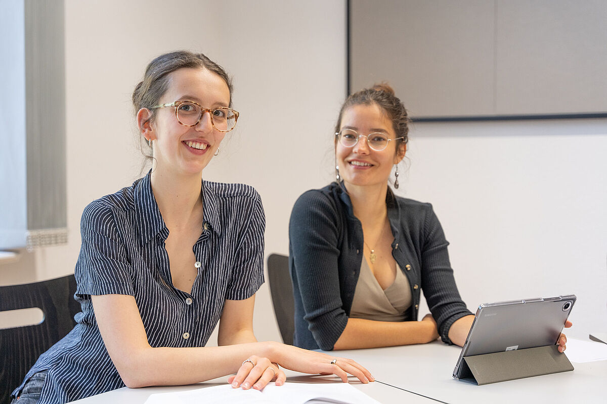 Zwei junge Frauen sitzen in einem hellen Arbeitsraum und schauen freundlich in die Kamera. Vor ihnen steht ein Tablet auf dem Tisch