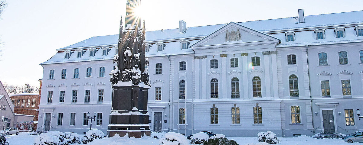 Das Hauptgebäude der Universität im Winter mit Schnee. Die Sonne scheint durchs Rubenowdenkmal. 