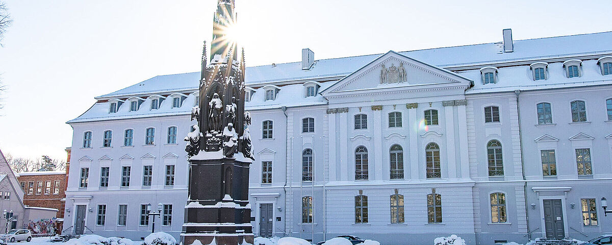 Das Hauptgebäude der Universität im Winter mit Schnee. Die Sonne scheint durchs Rubenowdenkmal. 