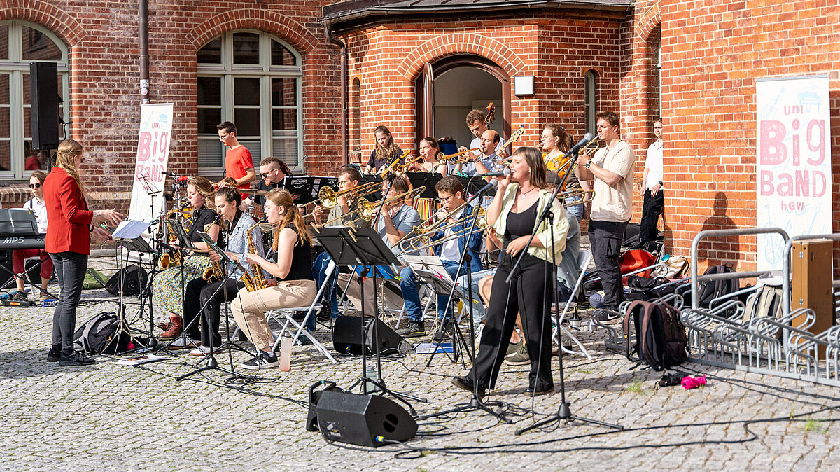 Eine größere Gruppe Musizierender sitzen mit Blasinstrumenten im Innenhof des Campus Beitzplatz und machen Musik.