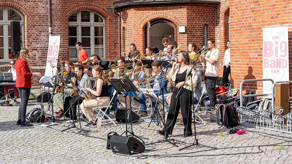 Eine größere Gruppe Musizierender sitzen mit Blasinstrumenten im Innenhof des Campus Lohmeyerplatz und machen Musik.